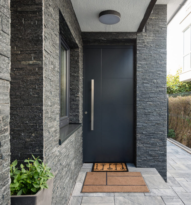 Anthracite aluminium entrance door with a long stainless pull handle, set in a recessed porch clad in dark stacked stone, with a covered soffit and ceiling light above the door